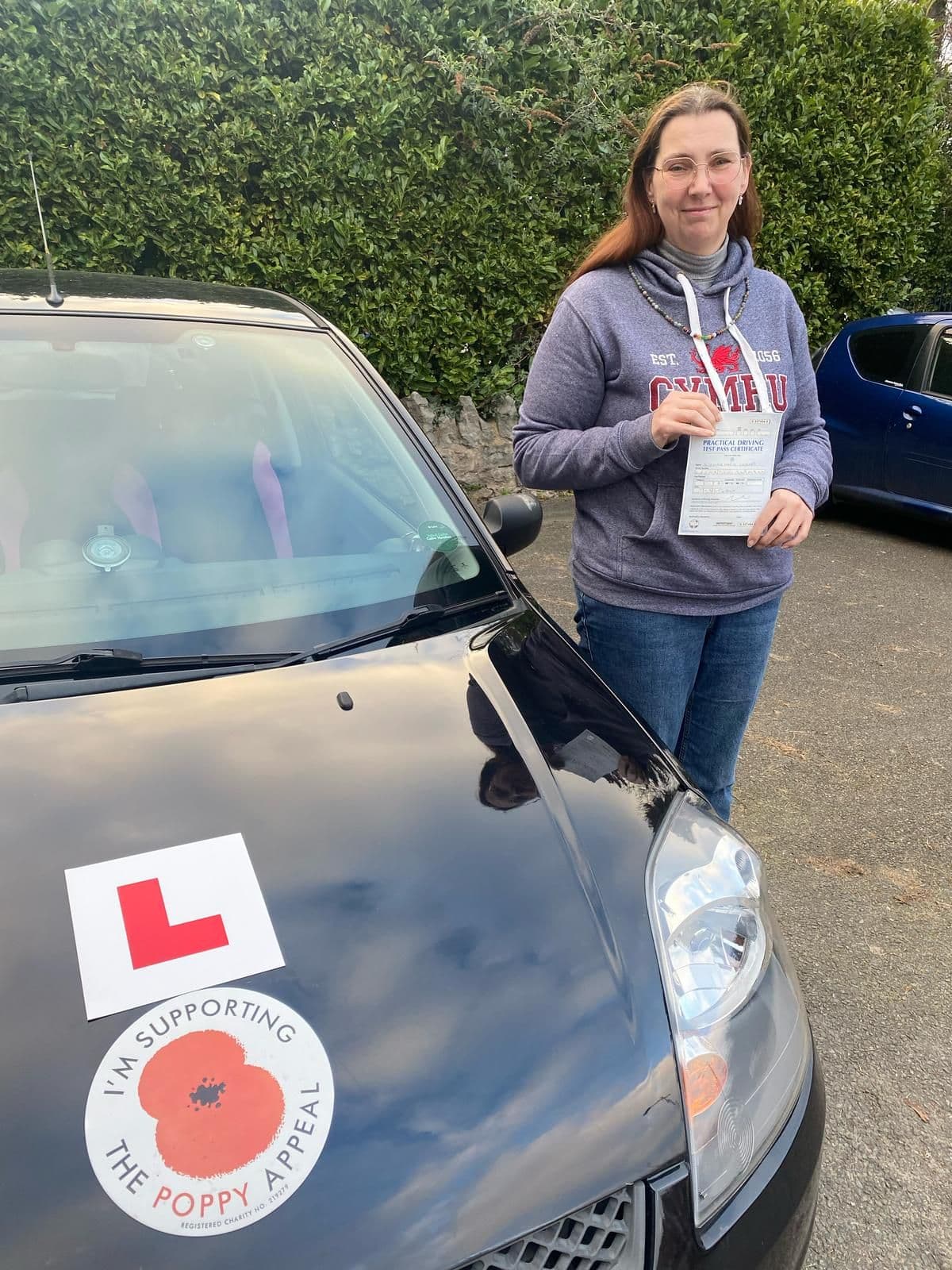 Woman holding a driving test pass certificate next to a black car with L plates.