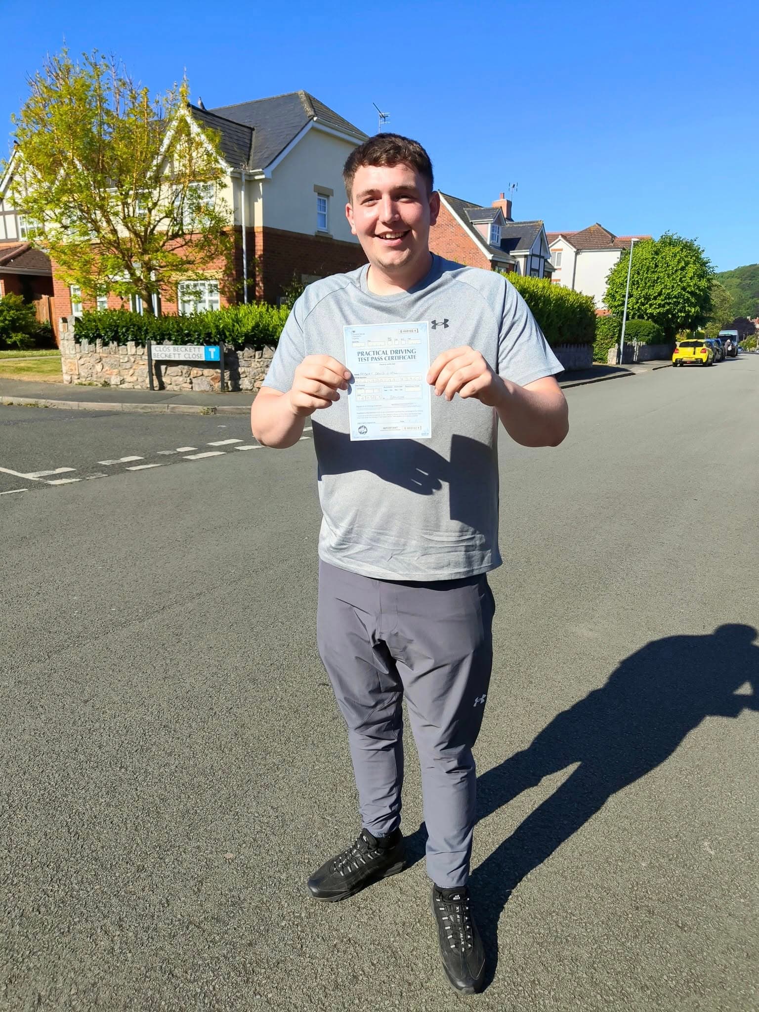 Smiling young man holding a practical driving test pass certificate on a sunny residential street.