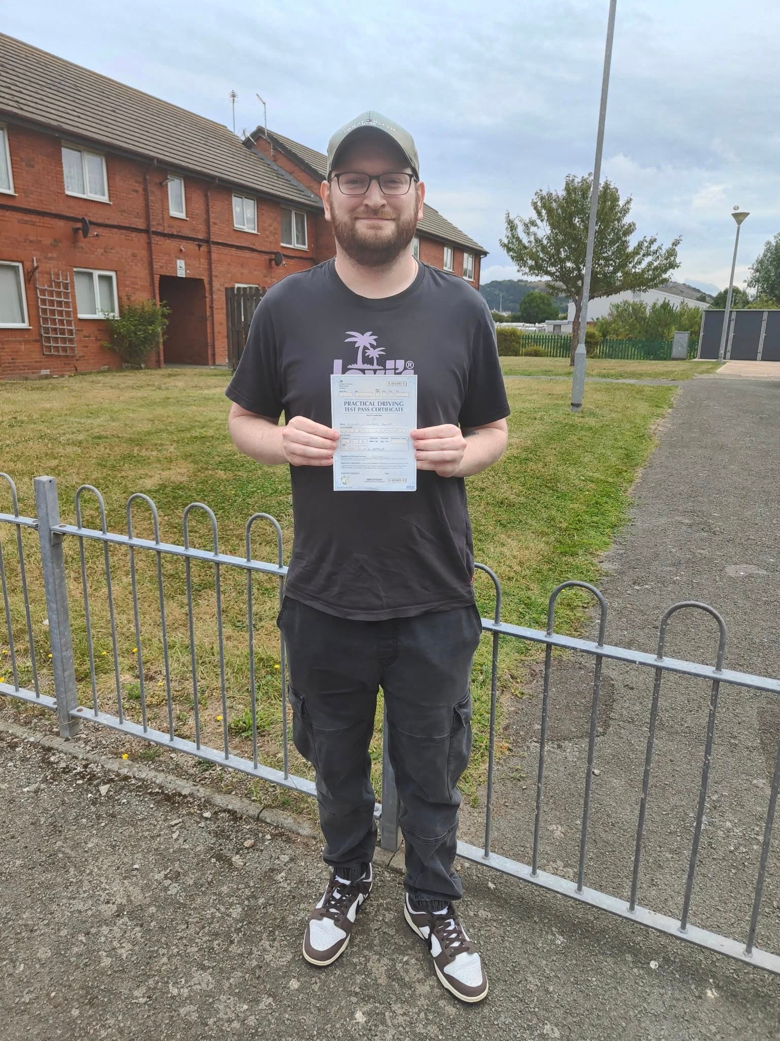 Smiling man in a cap holds up a practical driving test pass certificate outdoors.