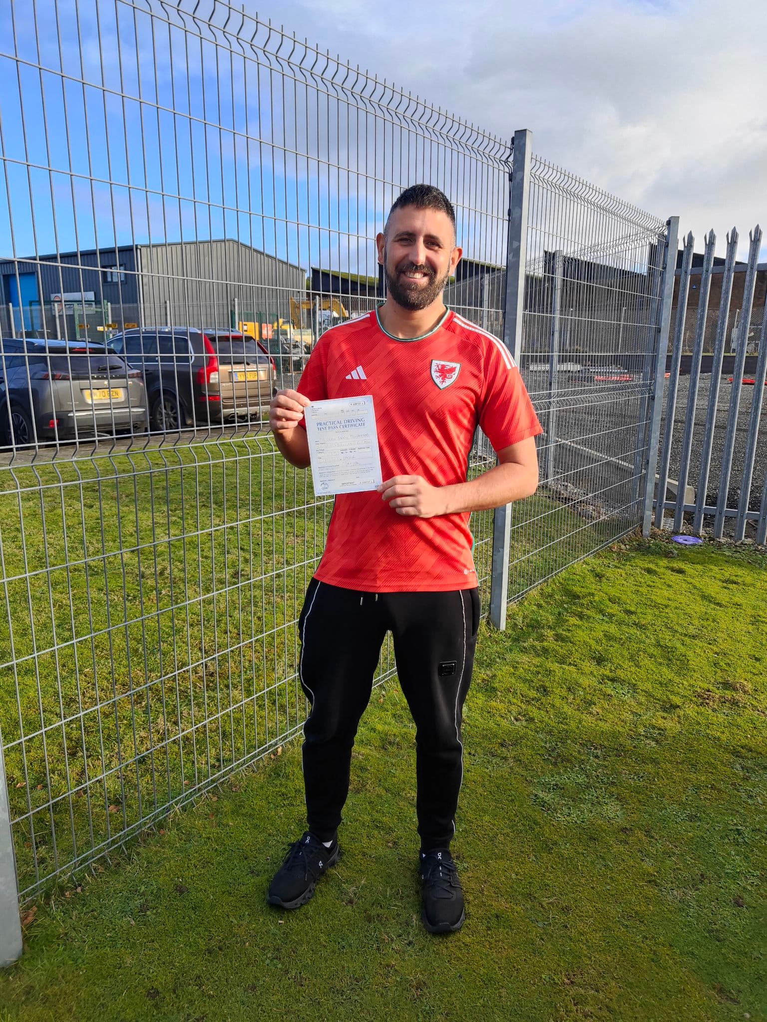 Smiling man in a red Wales football shirt holds a practical driving test pass certificate.